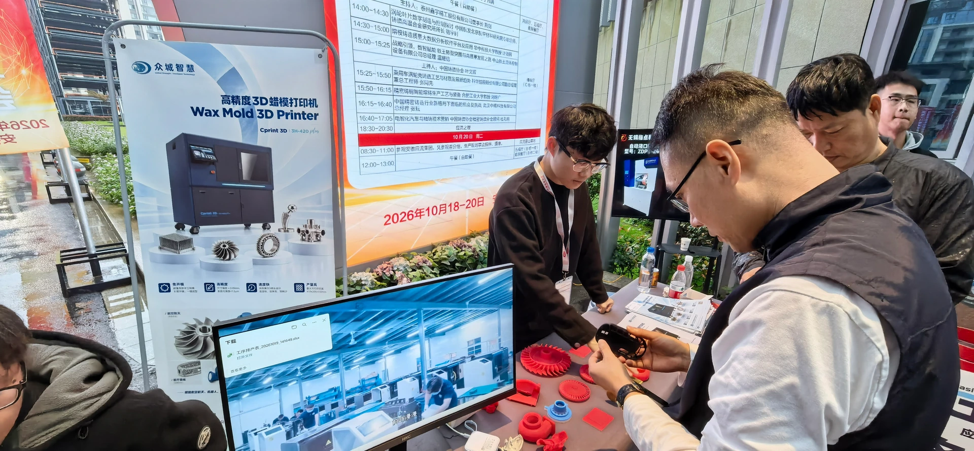 Des visiteurs examinent un échantillon près d’un support de présentation de l’imprimante cire Cprint 3D et de modèles en cire rouge à la conférence de fonderie